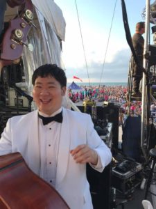 John (Bebo) Shiu is smiling at the camera while holding his bass. This photo was taken outside at a concert.