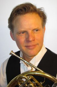 Headshot of Kevin Owen. He is smiling slightly, looking off camera while holding his horn.