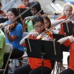 Young musicians from Boston String Academy, all wearing brightly colored shirts, are playing various string instruments. 