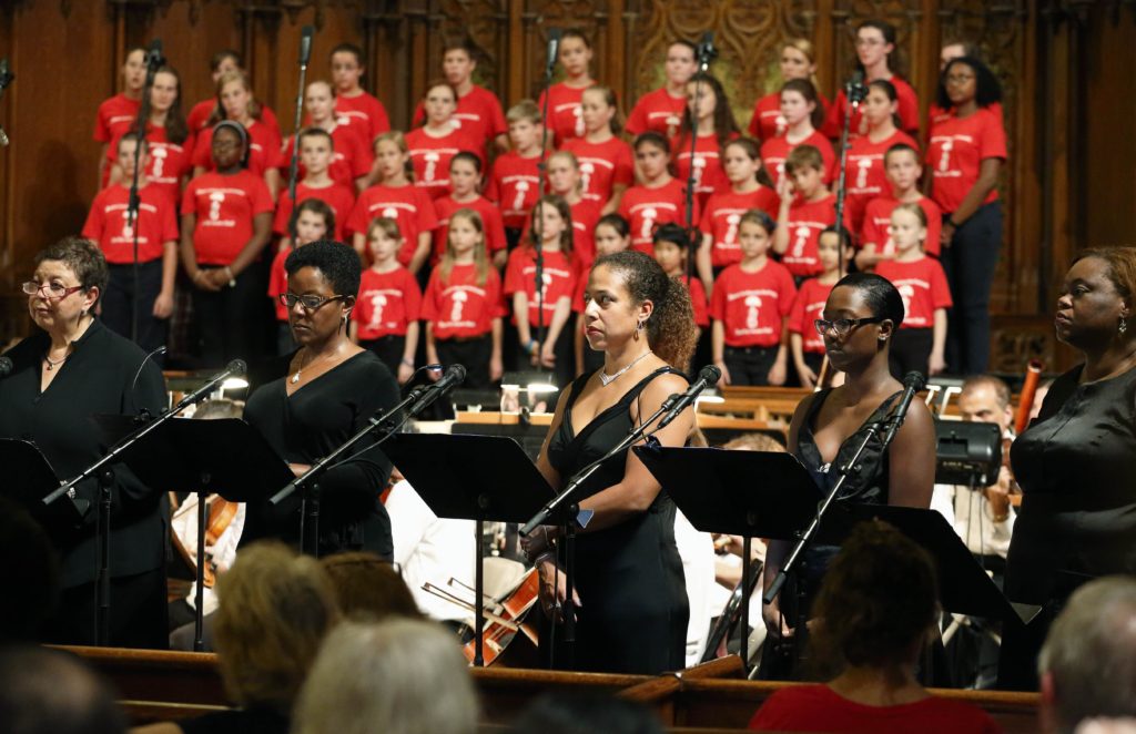Singers perform Griot Legacies by Trevor Weston. There are 5 singers upfront wearing black clothes, and around 30 singers behind them wearing red shirts.
