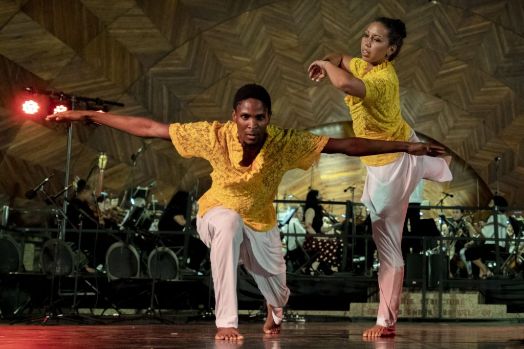 Performers of the piece Papa Loko by Gonzalo Grau onstage at the Hatch Shell. The dancers are wearing yellow shirts with white pants and have bare feet as they dance. The lower performer is bent down with their arms sticking out perpendicular to the ground.