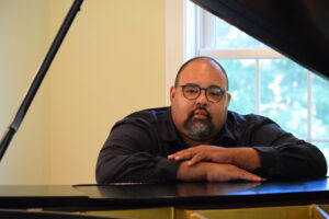 David Coleman has his arms rested on a piano. He is looking at the camera. 