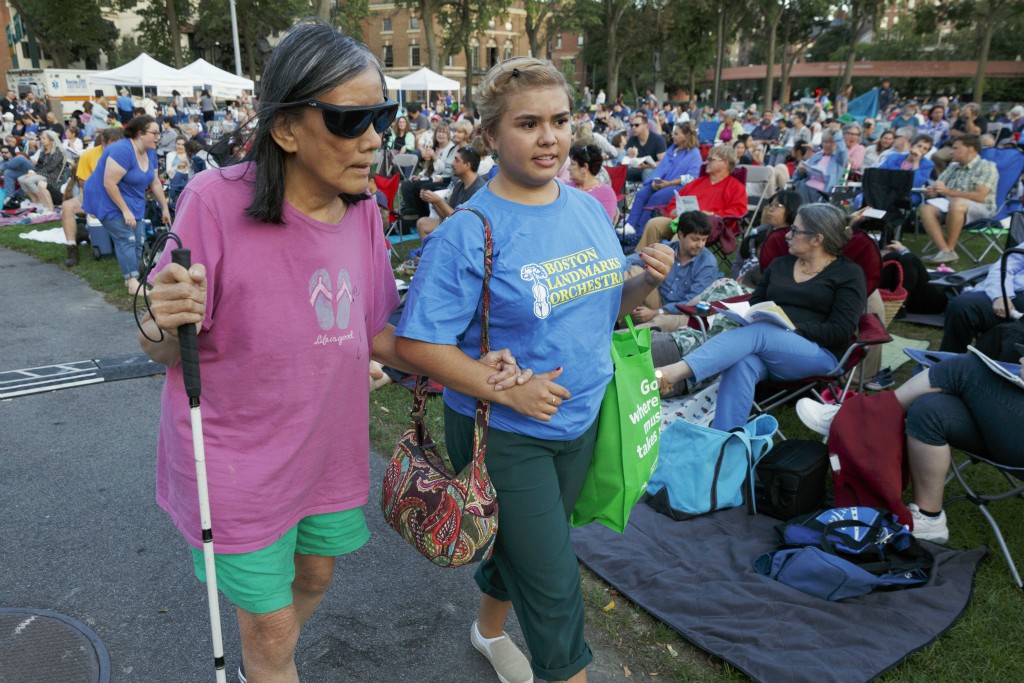 A staff member of Boston Landmarks Orchestra walk with a blind person with a guiding stick to assist them to their seat.