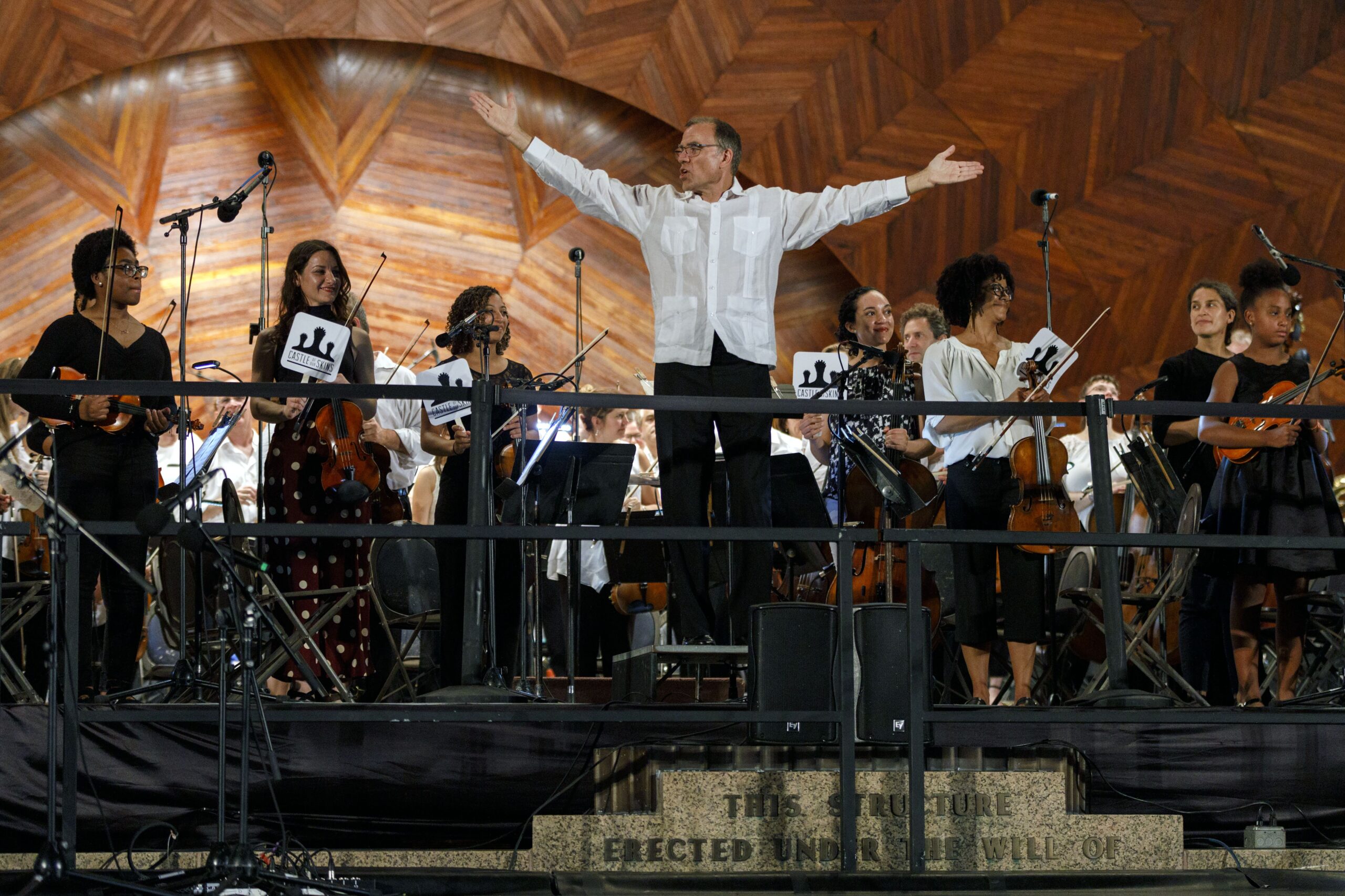 Music Director, Christopher Wilkins takes a bow, with his arms extended on either side of him, as members of Castle of our Skin stand behind him