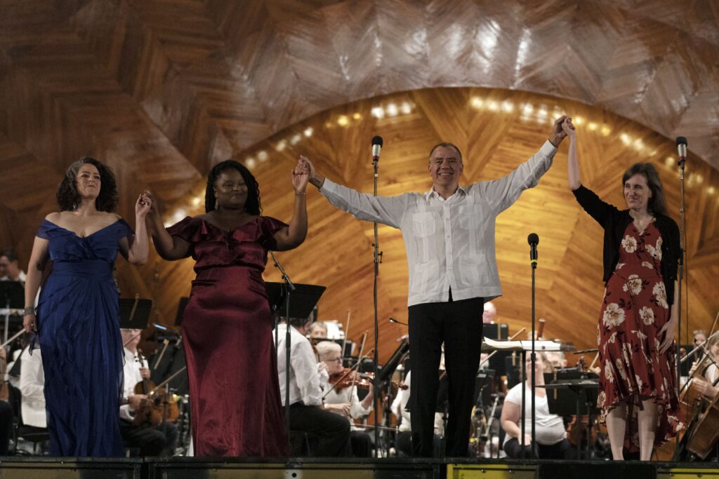 Christopher Wilkins, composer, Francine Trester, and singers Brianna Robinson and Carrie Cheron stand onstage at the Hatch Shell and raise their arms before taking a bow.