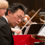 Dongsok Shin is looking to the side, as an orchestra plays behind him. 