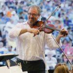 Greg Vitale plays the violin as a large crowd stands behind him, watching.