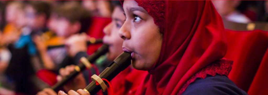 Child with a red hijab plays a clarinet; in the foreground are other children playing their instruments as part of the Link Up program.