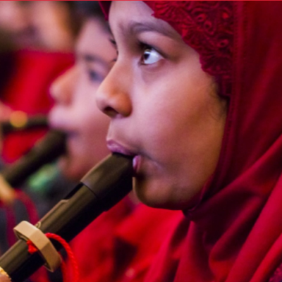 Child with a red hijab plays a clarinet; in the foreground are other children playing their instruments as part of the Link Up program.