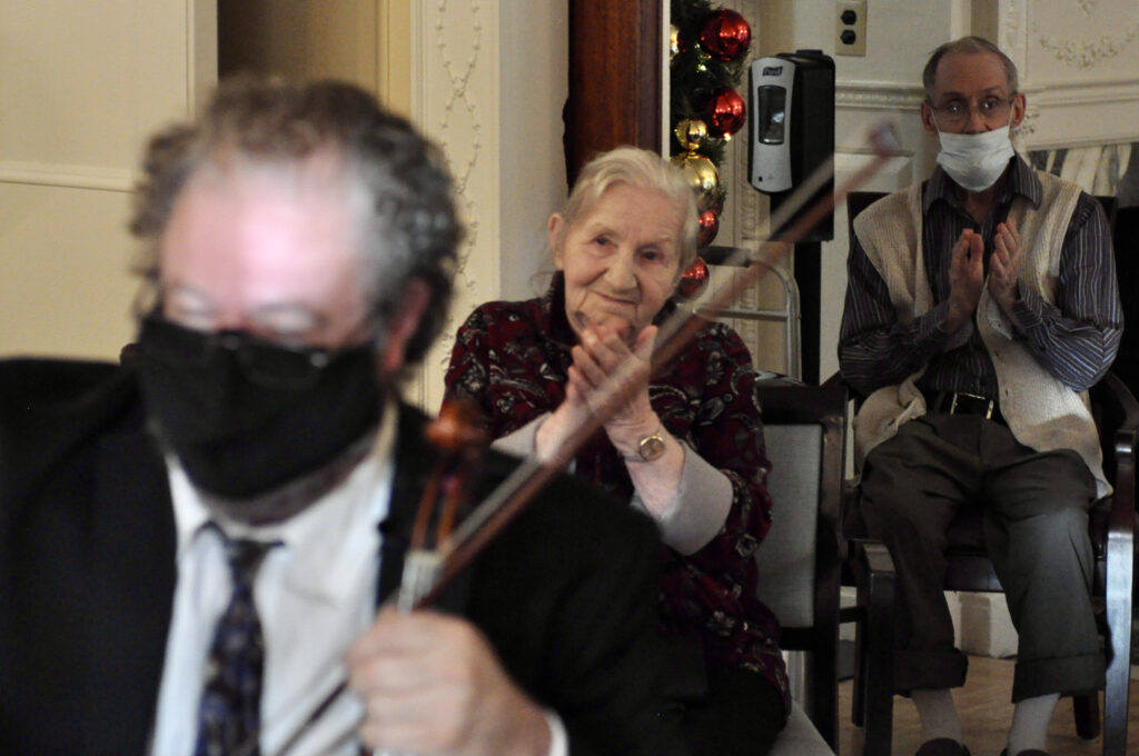 A string player holds a bow, while focused is a patron, smiling with her hands clasped together in applause. Another man behind her is wearing a mask and also applauding.