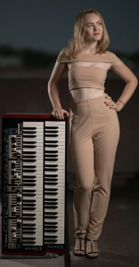 Anastassiya Petrova is standing next to her keyboard, wearing a beige top and pants, looking off to the side. The background is outside and appears to be evening.
