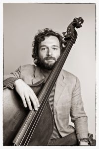 Max Ridley is sitting, holding his instrument. He stares straight at the camera.