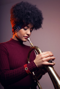 Milena Casado glances down at her horn, holding it. She is wearing a maroon sweater.