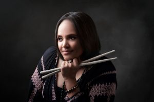 Headshot of Terri Lyne Carrington - She smiles, looking off to the side, holding two drumsticks.