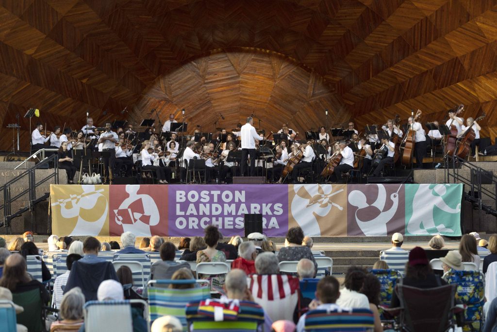 Hatch Shell stage with large banner of Landmarks Orchestra's new logo strewn across the stage. A large crowd watches from the lawn.